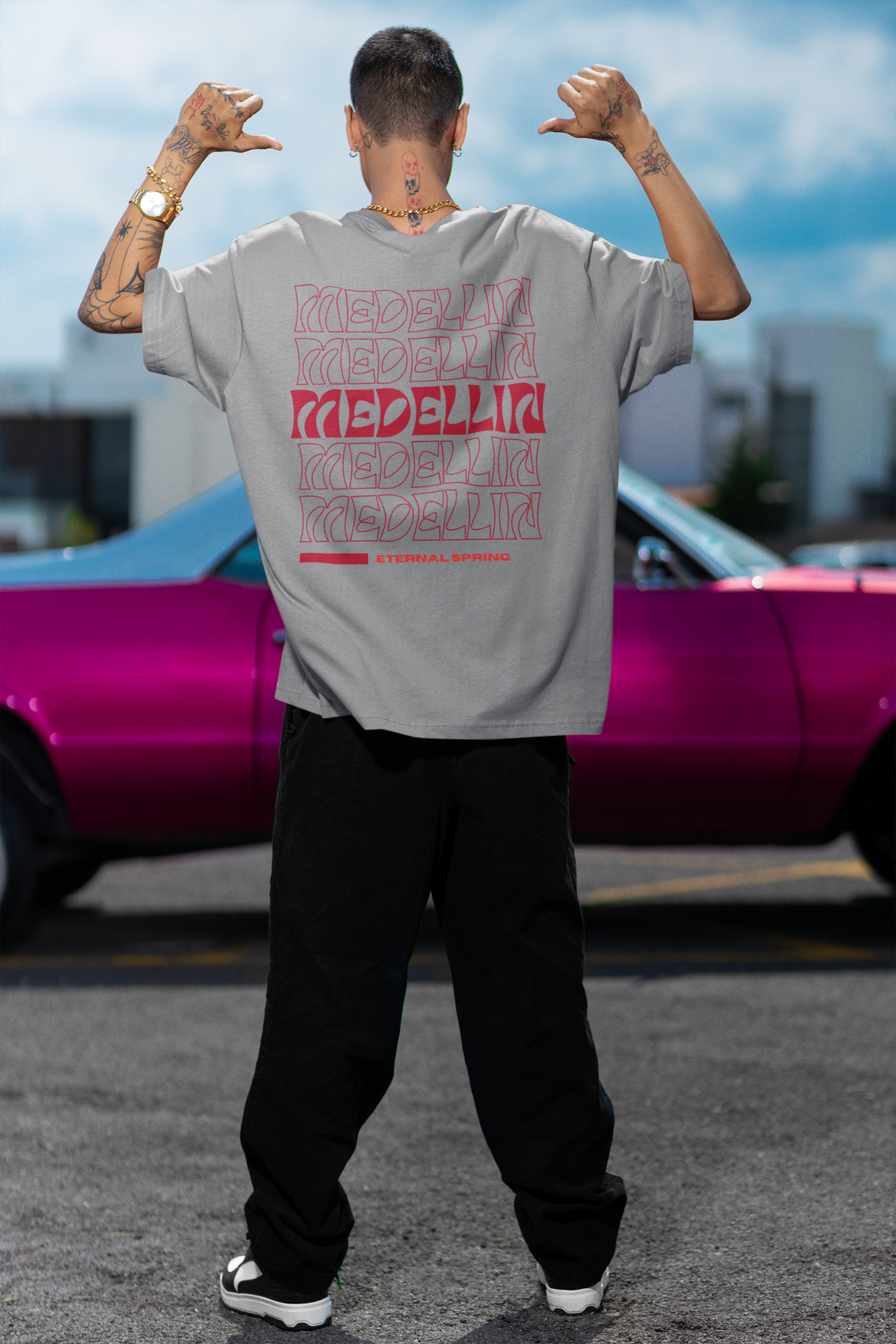 Back view of a male model wearing a grey T-shirt with red "MEDELLÍN" graphics, standing in front of a pink classic car in an urban setting.