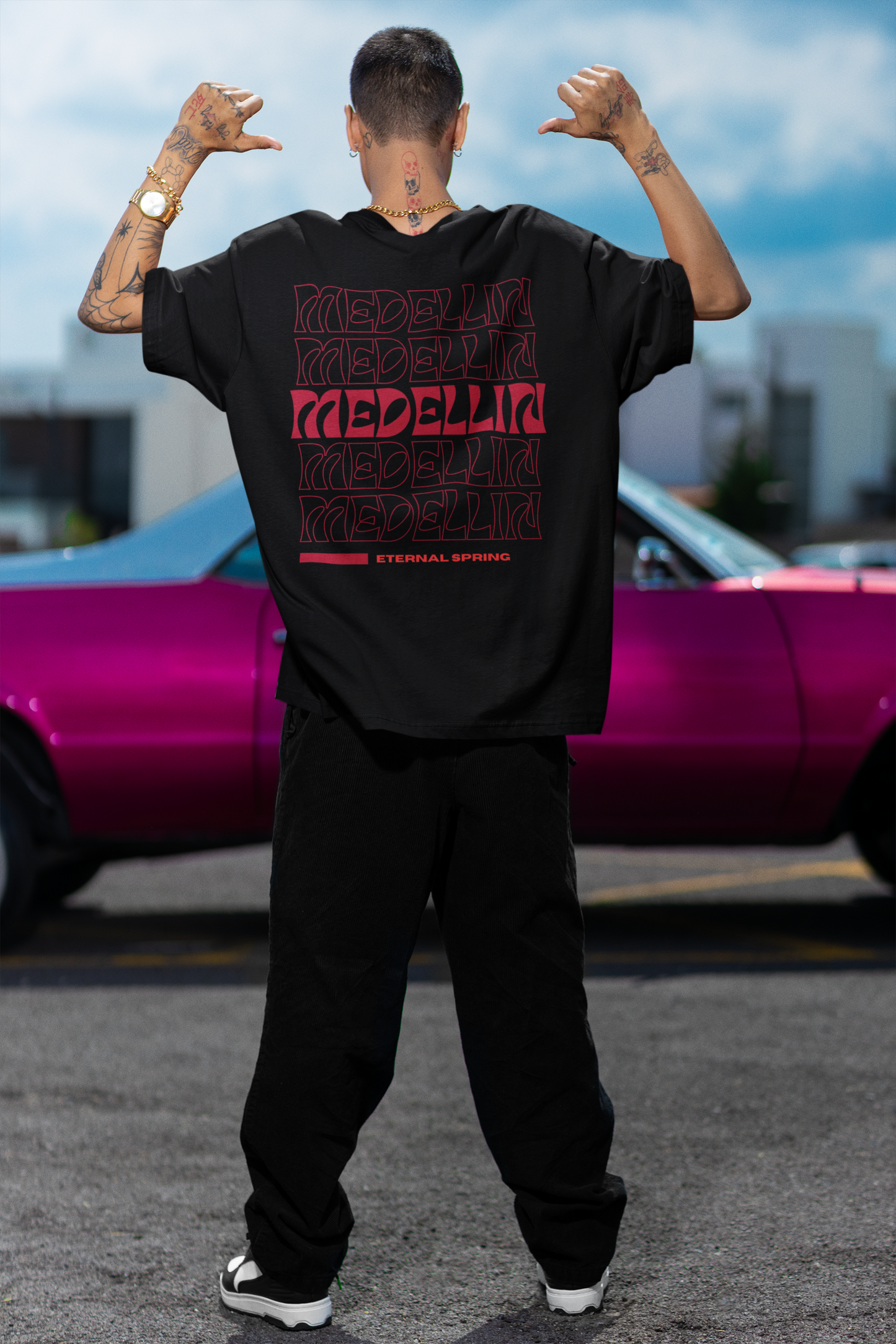Back view of a male model wearing a black T-shirt with red "MEDELLÍN" graphics, standing in front of a pink classic car in an urban setting.
