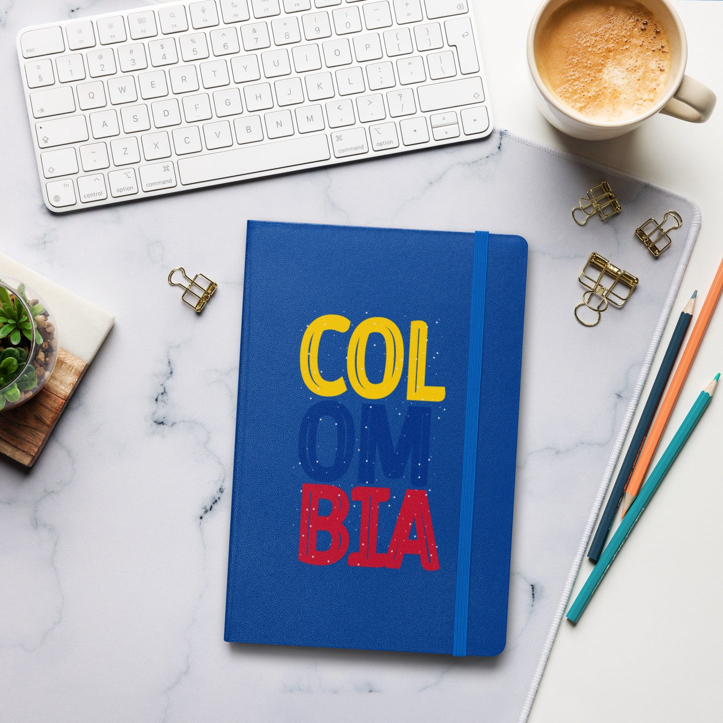 Blue hardcover notebook featuring bold 'COLOMBIA' text in yellow, blue, and red. Set on a stylish workspace with a keyboard, coffee, and stationery for a modern and creative vibe.