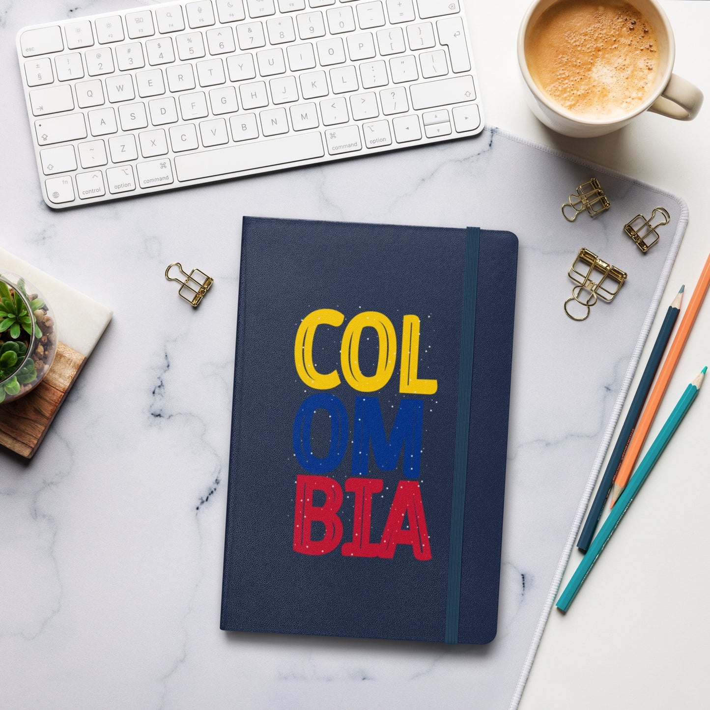 Navy hardcover notebook featuring bold 'COLOMBIA' text in yellow, blue, and red. Set on a stylish workspace with a keyboard, coffee, and stationery for a modern and creative vibe.