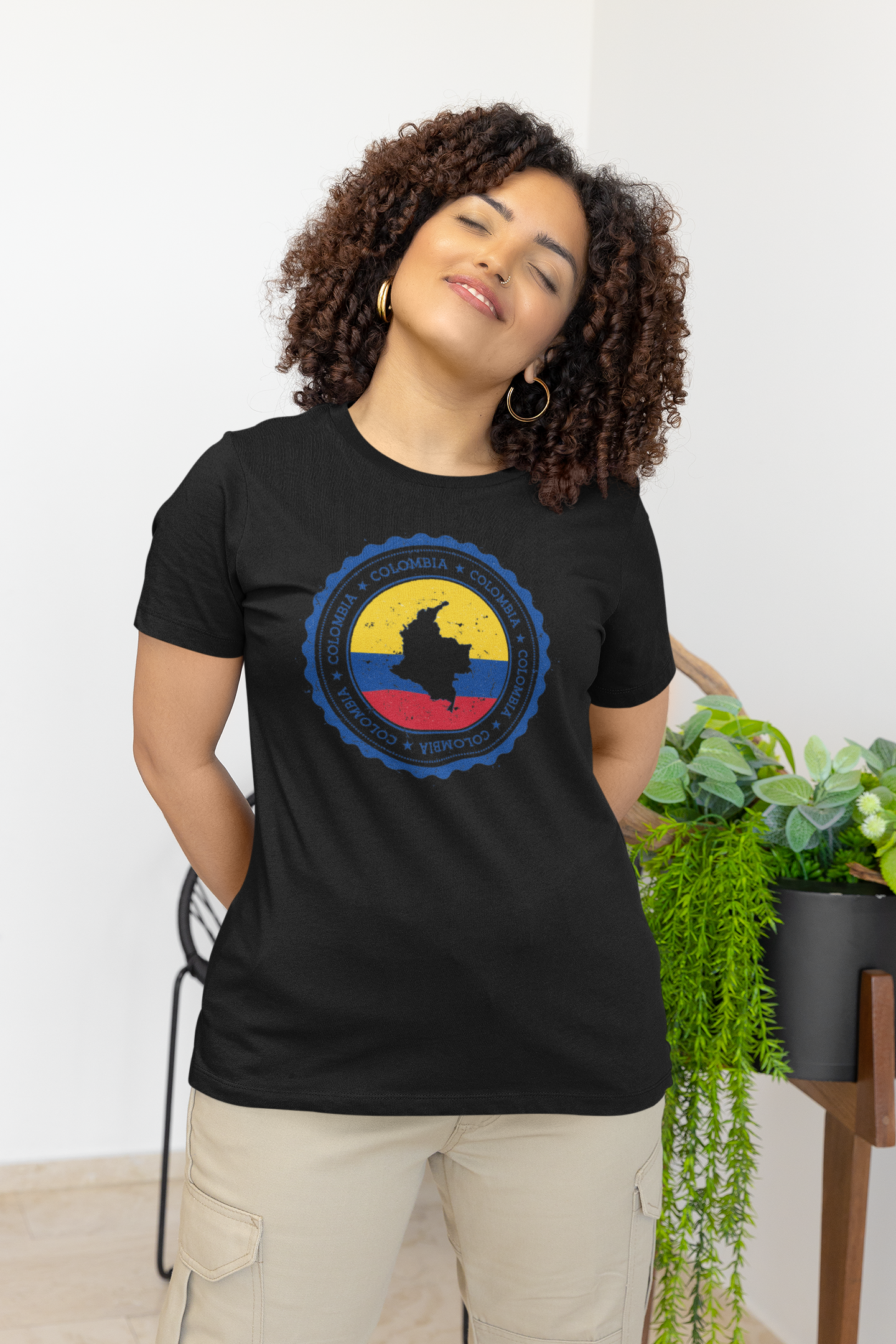 Curly-haired woman wearing a black T-shirt featuring a circular Colombian heritage design with yellow, blue, and red elements, standing confidently in a relaxed indoor setting.