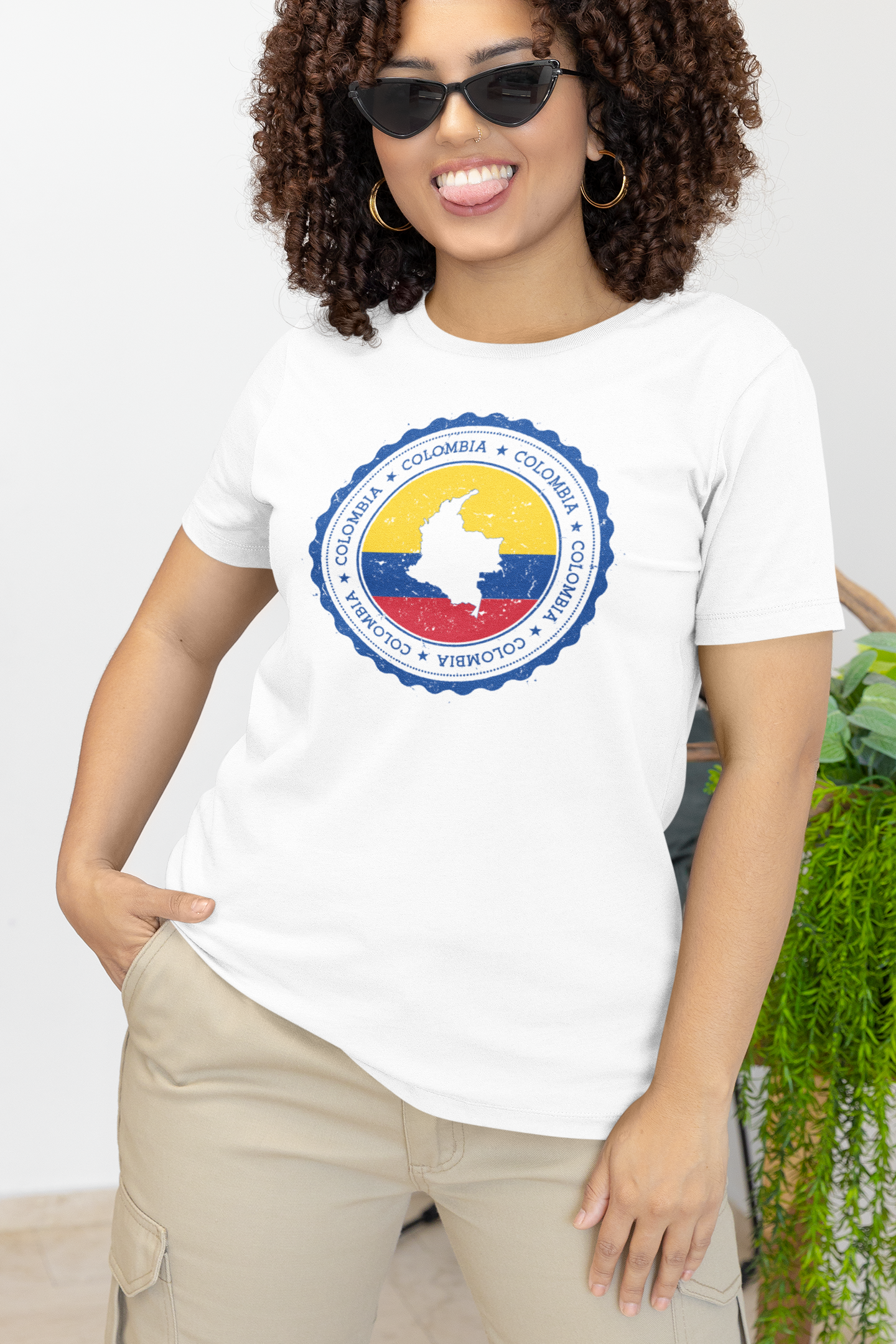 Smiling woman with curly hair wearing a white T-shirt featuring a circular Colombian heritage design with vibrant yellow, blue, and red accents, posing confidently indoors