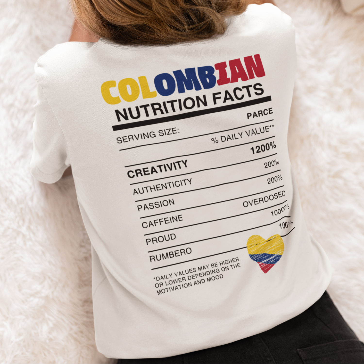 Back view of a woman lying face down on a cozy surface while reading, wearing a white T-shirt with a 'Colombian Nutrition Facts' design featuring colorful text and a heart-shaped Colombian flag graphic.