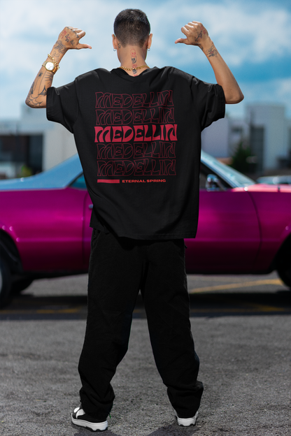 Back view of a male model wearing a black T-shirt with red "MEDELLÍN" graphics, standing in front of a pink classic car in an urban setting.