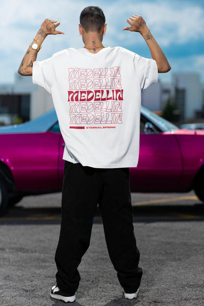 Back view of a male model wearing a white T-shirt with red "MEDELLÍN" graphics, standing in front of a pink classic car in an urban setting.