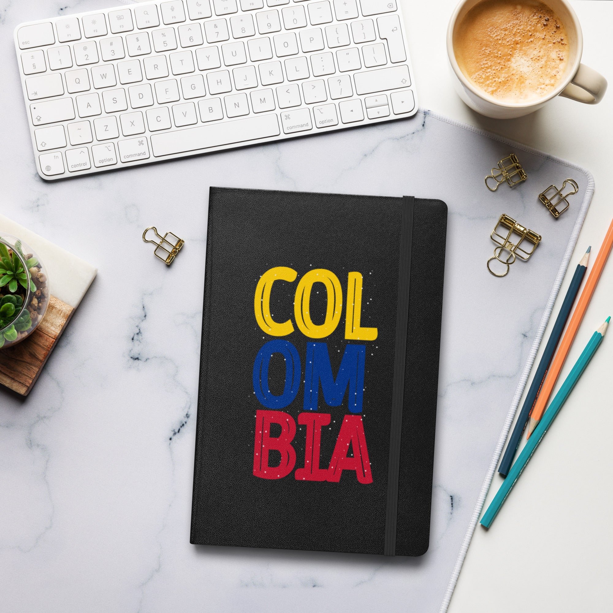 Black hardcover notebook featuring bold 'COLOMBIA' text in yellow, blue, and red. Set on a stylish workspace with a keyboard, coffee, and stationery for a modern and creative vibe."