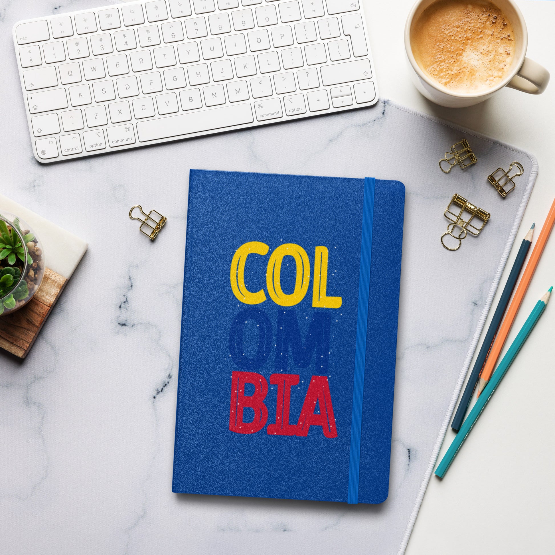 Blue hardcover notebook featuring bold 'COLOMBIA' text in yellow, blue, and red. Set on a stylish workspace with a keyboard, coffee, and stationery for a modern and creative vibe.