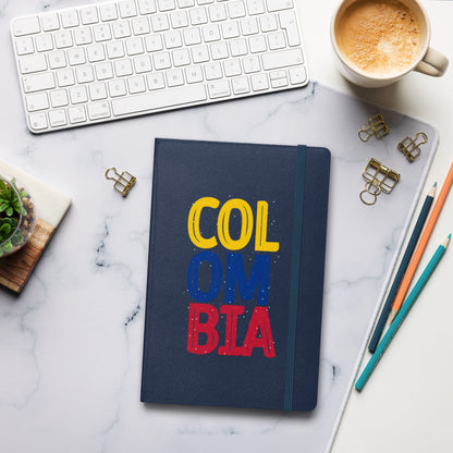 Navy hardcover notebook featuring bold 'COLOMBIA' text in yellow, blue, and red. Set on a stylish workspace with a keyboard, coffee, and stationery for a modern and creative vibe.