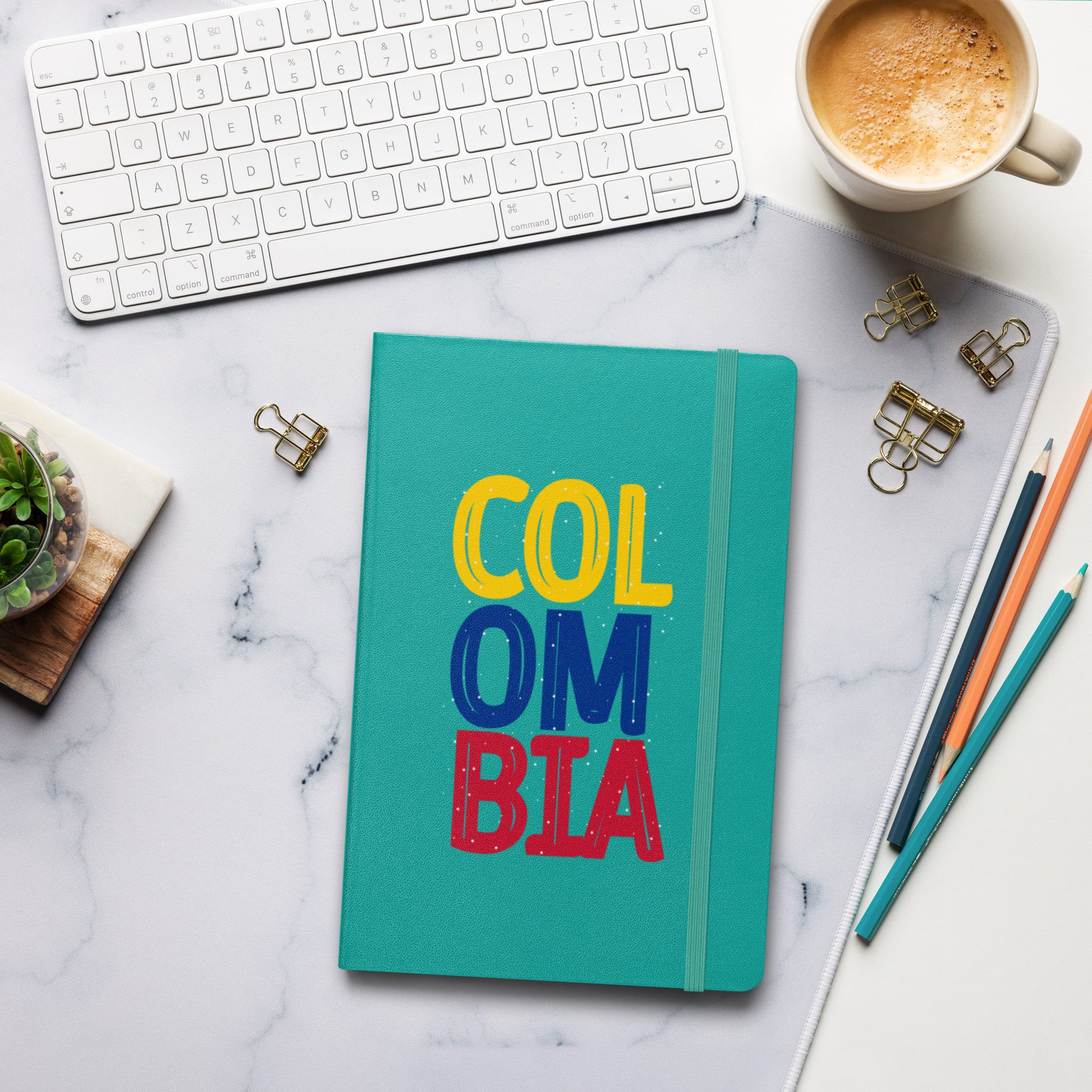 Turquoise hardcover notebook featuring bold 'COLOMBIA' text in yellow, blue, and red. Set on a stylish workspace with a keyboard, coffee, and stationery for a modern and creative vibe.