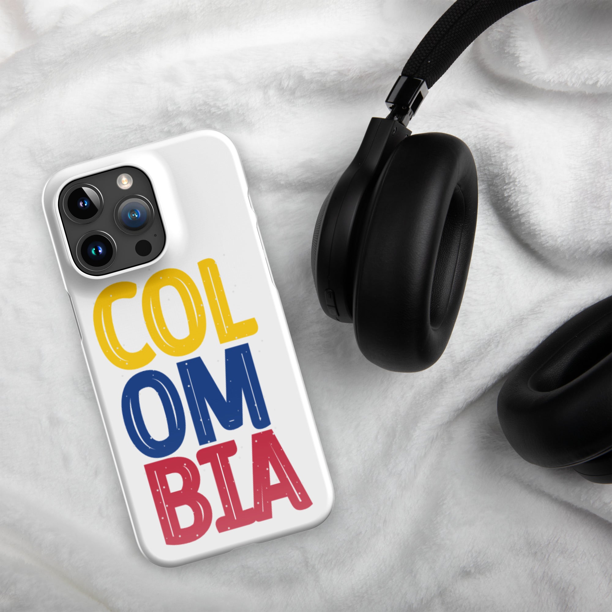 Glossy white iPhone 15 Pro Max case featuring a bold and colorful 'COLOMBIA' design in yellow, blue, and red. Displayed on a soft white fabric background next to black over-ear headphones, perfect for showcasing Colombian pride.